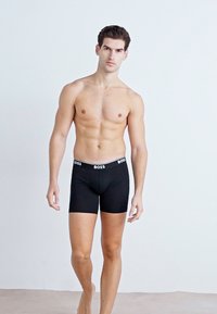 Schwarze Boxer-Brief mit einem weißen Bund, der das "BOSS"-Logo trägt. Weicher Stoff, körpernahe Passform, mittellange Oberschenkel-Länge, glatte Textur.