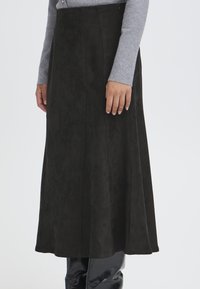 Gonna maxi in suede nera con una texture liscia, caratterizzata da cuciture verticali e un fondo svasato. Indossata con un top grigio a maniche lunghe.