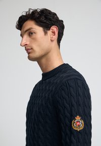 Mörkblå kabelstickad tröja med ribbad rund halsringning. Har ett texturerat mönster och en rund broderad märke på vänster arm.