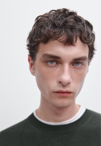 Gebreide trui in donkergroen, gedragen over een wit T-shirt met ronde hals. Heldere blauwe ogen, lichte huid en kort, krullend haar met textuur. Neutrale achtergrond.