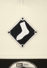 Cream-colored cap with a diamond-shaped black patch featuring a white sock embroidery. Brand label at the bottom. Smooth texture.