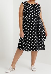 Vestido negro con lunares, con corsé ajustado, falda acampanada y cinturón atado. Combinado con zapatos blancos de slip-on. Tejido texturizado y escote redondo.
