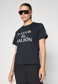 T-shirt in cotone nero con la scritta "le CLUB du MALBÔN" in grande carattere bianco. Abbinato a pantaloni sportivi neri, il modello indossa occhiali da sole scuri.