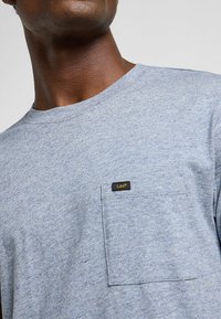 T-shirt grigio a maniche corte realizzato in un tessuto texturizzato. Presenta una tasca sul petto sinistro e un piccolo etichetta con il logo "Lee" in nero e oro.