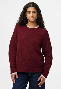 Femme avec de longs cheveux bouclés portant un pull en maille bordeaux avec des bordures blanches et un jean bleu foncé, se tenant devant un fond blanc uni.