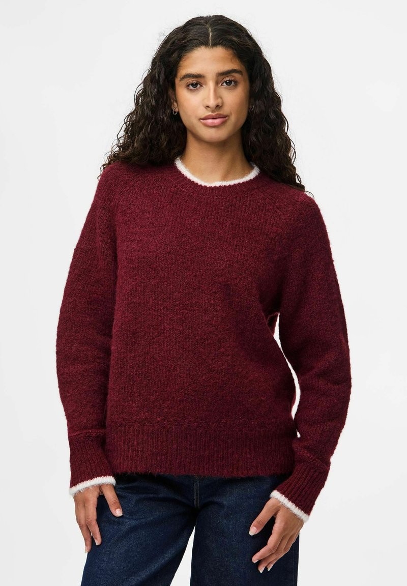 Femme avec de longs cheveux bouclés portant un pull en maille bordeaux avec des bordures blanches et un jean bleu foncé, se tenant devant un fond blanc uni.