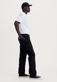 Zwarte denim broek met een rechte fit, wit t-shirt en zwarte pet. Details omvatten zijzakken en klassieke sneakers.