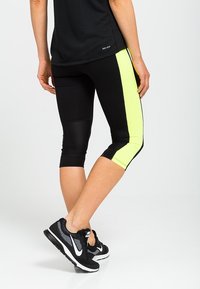 Osoba ubrana w czarne legginsy do kostek w neonowo-żółtym kolorze i czarne buty do biegania Nike, stojąca z jedną nogą skrzyżowaną za drugą.