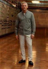 Szary button-up koszula, białe spodnie i czarne buty, stojący na wypolerowanej drewnianej podłodze z vintage'owym industrialnym tłem.