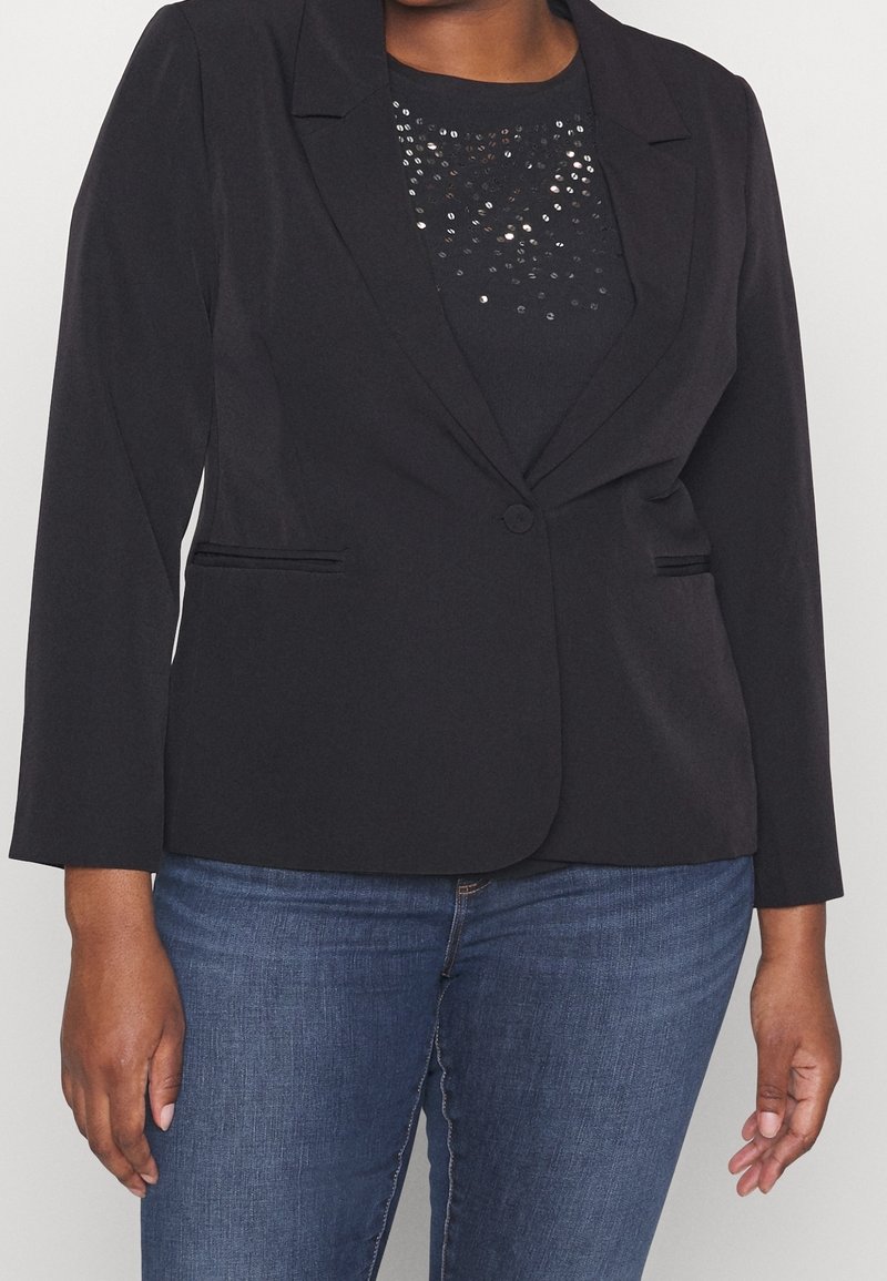 Femme portant un blazer noir ajusté sur un haut noir pailleté, associé à un jean bleu moyen, debout les bras détendus le long du corps.