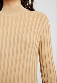 Pull en maille côtelée beige avec des rayures verticales et un col montant ajusté.