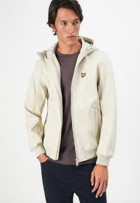Uomo che indossa una giacca a zip beige con cappuccio sopra una maglia grigia scuro e pantaloni blu navy, in piedi con una mano in tasca su uno sfondo bianco.