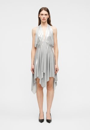 Kobieta ubrana w bezrękawnikową, srebrną metaliczną sukienkę halter z asymetrycznym obszyciem w kształcie chusteczki, w połączeniu z czarnymi czółenkami na szpic.