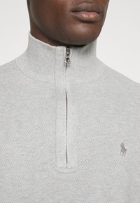 Ljusgrå ziphoodie med texturerad stickad design, hög krage och liten broderad logotyp på bröstet. Silverfärgad dragkedja.