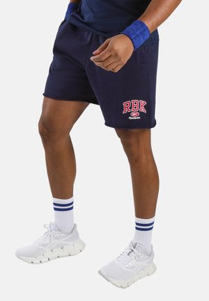 Mann trägt marineblaue Reebok-Shorts mit RBK-Logo, weiße Sportschuhe, weiße Socken mit blauen Streifen und blaue Handgelenkbänder, steht auf weißem Hintergrund.