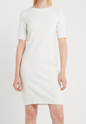 Femme portant une robe blanche côtelée, ajustée, à manches courtes et longueur genou, sur un fond clair uni.