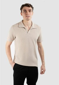 Kortärmad beige polo med ribbad textur, öppen krage och rak nederkant. Bärs med svarta byxor, visas mot en grå bakgrund.