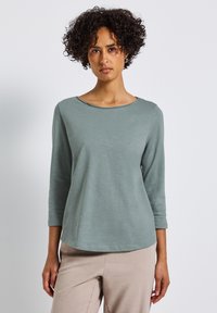 Helles grünes, langärmliges Oberteil mit einem Bootsausschnitt; aus weichem, strukturiertem Stoff gefertigt. Das Design zeichnet sich durch eine lockere Passform und einen subtilen Glanz aus.