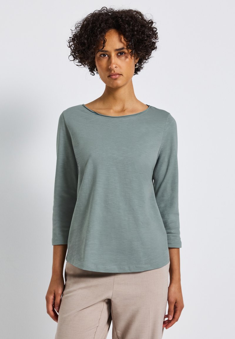 Helles grünes, langärmliges Oberteil mit einem Bootsausschnitt; aus weichem, strukturiertem Stoff gefertigt. Das Design zeichnet sich durch eine lockere Passform und einen subtilen Glanz aus.