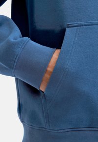 Sudadera azul con puños acanalados, que cuenta con un bolsillo canguro. La tela parece suave y con textura, con costuras visibles que destacan.