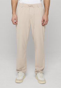 Pantalon de jogging beige avec une taille élastique et un cordon de serrage. Tissu lisse, coupe décontractée, poches latérales, associé à des baskets blanches.
