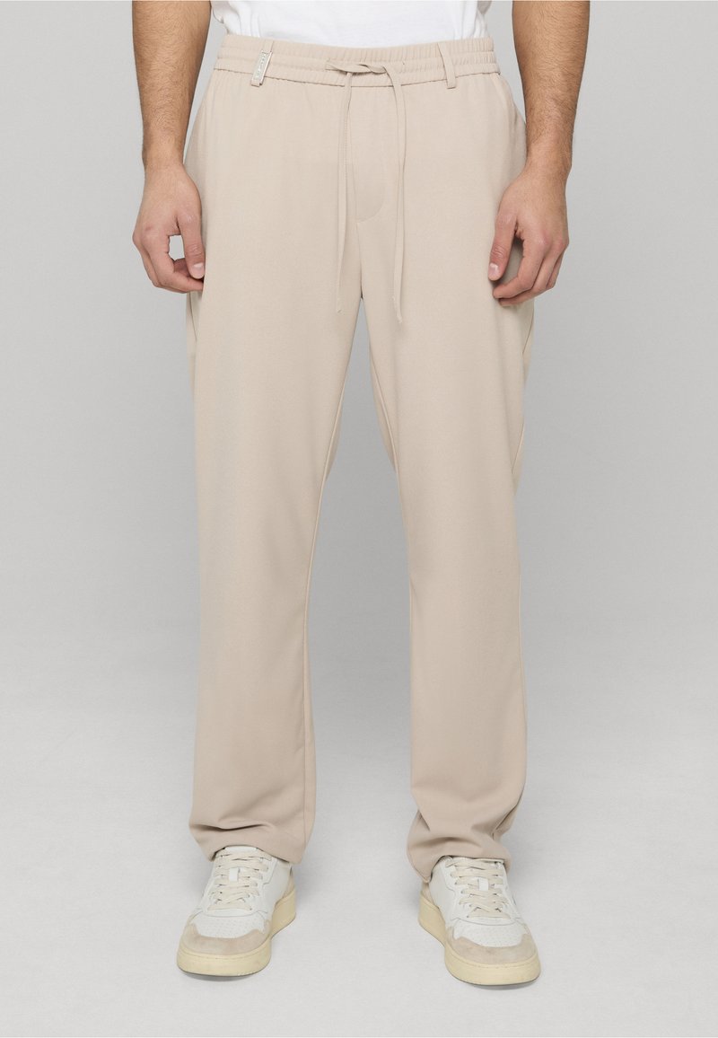 Pantalon de jogging beige avec une taille élastique et un cordon de serrage. Tissu lisse, coupe décontractée, poches latérales, associé à des baskets blanches.