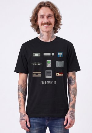 Man with tattoos and wavy hair smiles wearing a black shirt with retro gaming consoles and "I'm lovin' it" text.