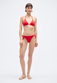 Set bikini rosso con top a triangolo con sottili spalline e slip con laccetti ai lati. Realizzato in materiale liscio ed elastico.