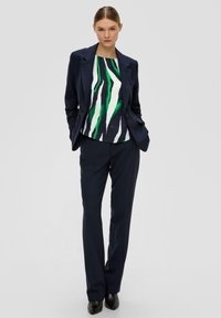 Marineblauer maßgeschneiderter Blazer über einem gemusterten Oberteil mit grünen und weißen Streifen, kombiniert mit dunklen Hosen und schwarzen spitzen Schuhen.