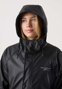 Czarna wodoodporna kurtka z kapturem, zatrzaskami, regulowanymi ściągaczami oraz logo na piersi. Gładka faktura i elegancki design.