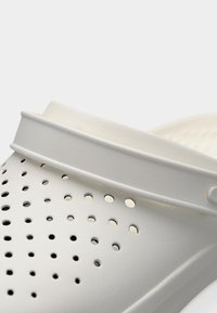 Λευκό παντόφλα με τρύπες και ρυθμιζόμενο λουράκι στη φτέρνα, κατασκευασμένη από ελαφρύ υλικό, εμφανιζόμενη σε κοντινή λήψη σε απλό φόντο.