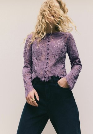 Personne blonde aux cheveux bouclés porte un haut en dentelle violette à manches longues et un pantalon foncé, posant avec une main dans la poche devant un fond uni.