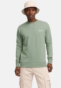 Sudadera verde claro con puños y dobladillo acanalados. Presenta un sutil logo en el pecho, combinada con pantalones cargo beige y un sombrero de cubo beige.