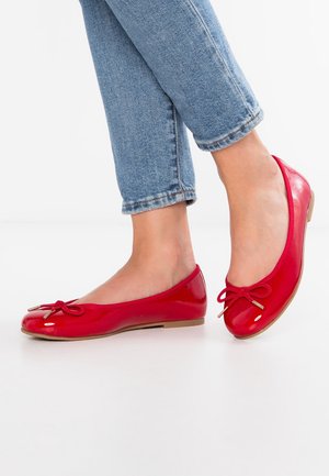 Jambes en jean bleu portant des ballerines rouges brillantes avec de petits nœuds, debout sur une surface blanche.