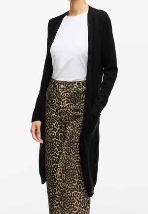 Vrouw die een wit T-shirt, een lange zwarte cardigan en een broek met hooggesneden luipaardprint draagt, staand met één hand in haar zak.