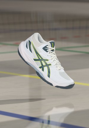 Λευκό και γκρι αθλητικό παπούτσι Asics με νεον κίτρινα στοιχεία να αιωρείται πάνω από το δάπεδο ενός γυμναστηρίου που έχει σημαδευτεί με πολύχρωμες γραμμές γηπέδου.