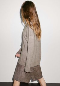 Beige gestrickter Pullover mit lockerer Passform und langen Ärmeln, kombiniert mit braunen Wildledershorts. Das Modell hat lange, wellige Haare und blickt von der Kamera weg.