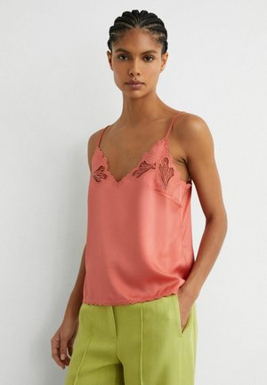 Femeie cu părul împletit, purtând o camisă de satin roz coral cu dantelă și pantaloni verde deschis, stând cu o mână în buzunar.