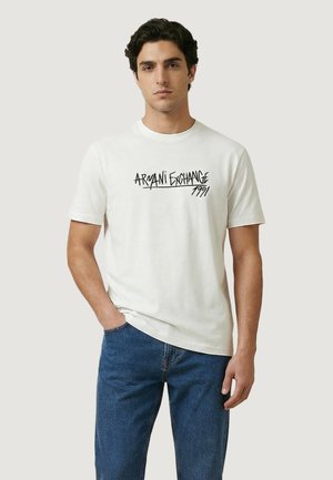 Mann trägt weißes Armani Exchange 1991 Grafik-T-Shirt und blaue Jeans, steht mit einer Hand in der Tasche vor einfachem Hintergrund.