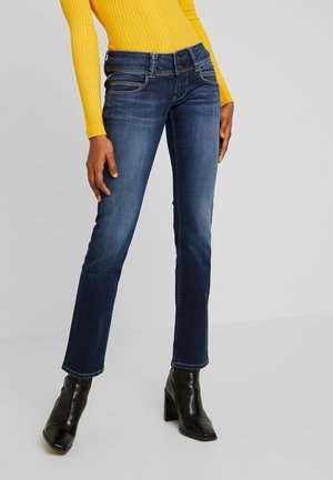 Femme portant un jean slim bleu marine, un haut côtelé à manches longues jaune et des bottines à talons noires, debout sur un sol gris.