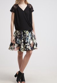 Top nero con maniche in pizzo, abbinato a una gonna nera floreale con motivi colorati. La modella indossa stivali con tacco aperti neri.