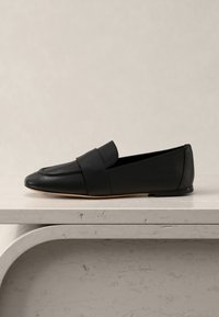 Μαύρο δερμάτινο φλατ μοκασίνι με φαρδιά λουρίδα στο πάνω μέρος, τοποθετημένο σε ραφιέρα με ανοιχτόχρωμη υφή πάνω σε ουδέτερο φόντο.