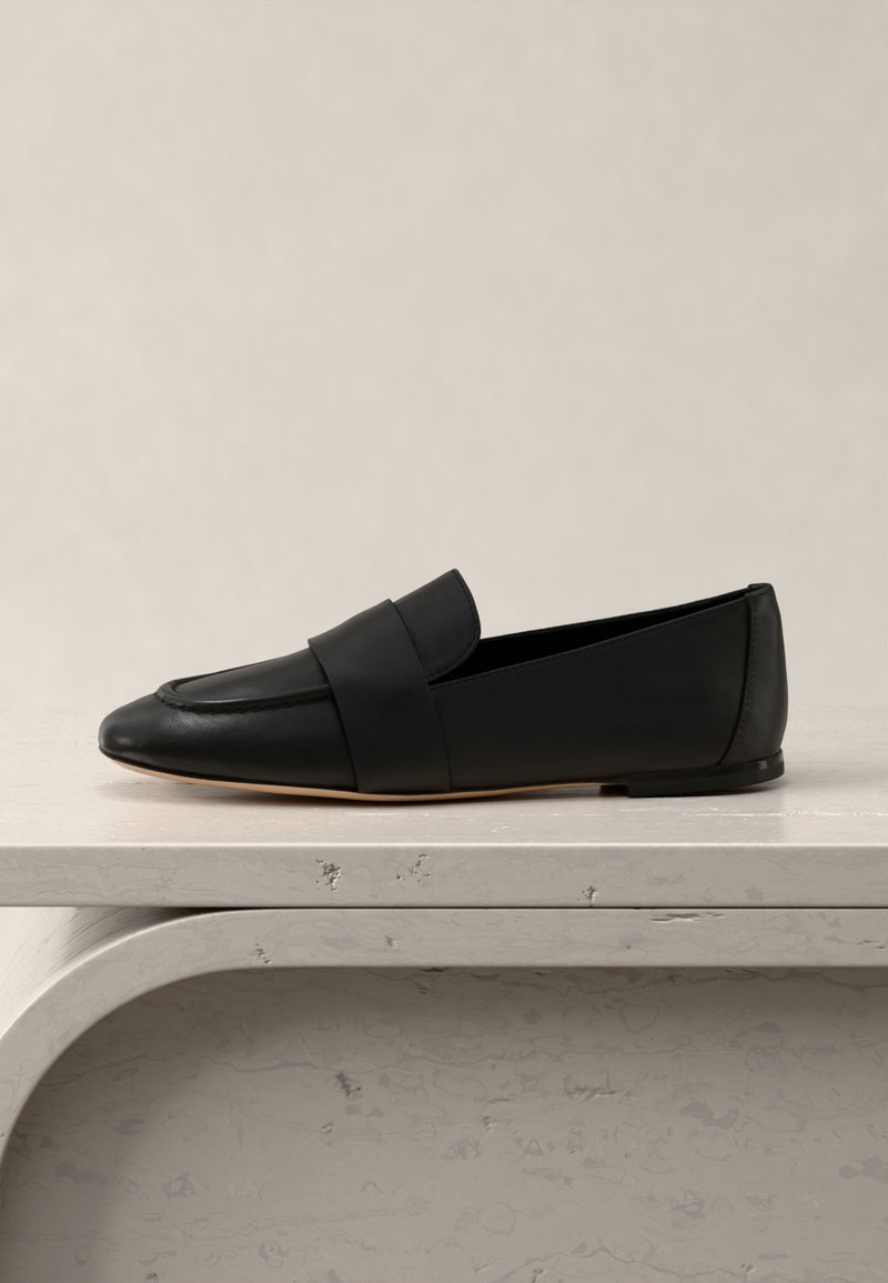 Μαύρο δερμάτινο φλατ μοκασίνι με φαρδιά λουρίδα στο πάνω μέρος, τοποθετημένο σε ραφιέρα με ανοιχτόχρωμη υφή πάνω σε ουδέτερο φόντο.