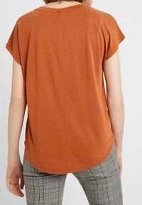 Rust orange short-sleeve t-shirt, soft fabric, rounded neckline, slightly loose fit, back seam detail, worn with checked trousers.