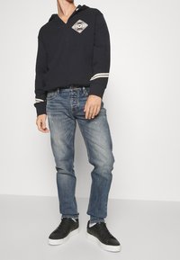 Marinblå huvtröja med vita detaljer, prydd med en diamantgrafik på bröstet; ihop med urblekta blå jeans och svarta sneakers.
