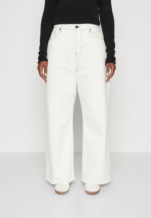 Jeans bianchi a gamba larga con cuciture a contrasto, vita alta e orlo sfilacciato, abbinati a un top aderente nero a maniche lunghe e sneakers bianche.