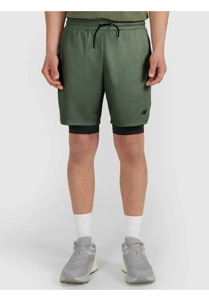 Dolna część ciała osoby noszącej zielone sportowe spodenki z czarnymi legginsami kompresyjnymi pod spodem, białe skarpetki i jasnoszare buty do biegania.