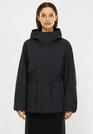 Kvinde iført en sort hættejakke med forlommer og justerbare snore, stående mod en ensartet lys baggrund.