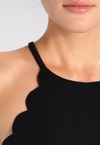 Musta hihaton toppi, jossa on aaltoileva pääntie, valmistettu teksturoidusta kankaasta, jossa on sileä pinta, sekä säädettävät hihnat.