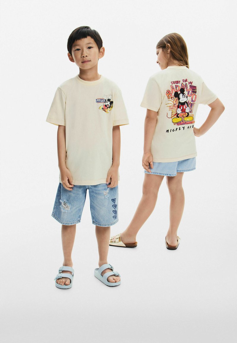 T-maica boje kreme s grafikom Mickey Mouse na prednjoj i stražnjoj strani, uparena s izlizanim trapericama, svijetloplavim sandalama i zlatnim kliznim sandalama.